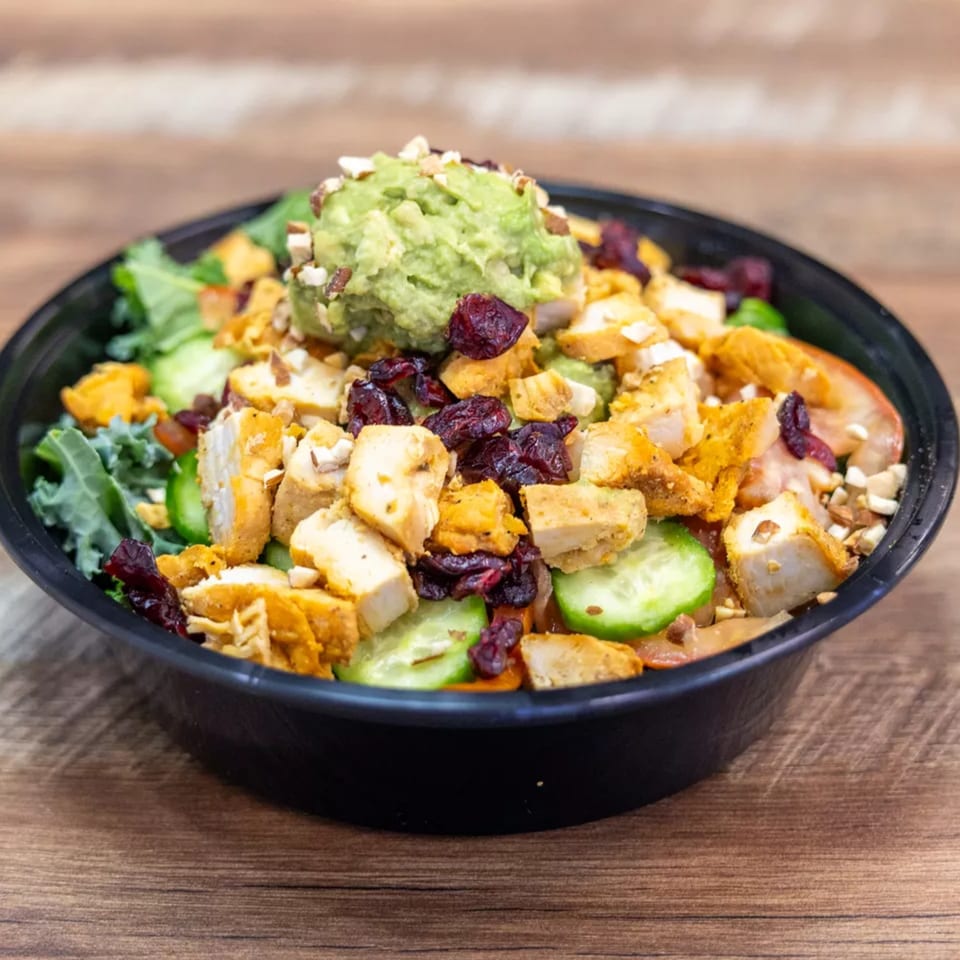 A vibrant salad with chicken, cranberries, cucumbers, and guacamole in a black bowl on a wooden table.