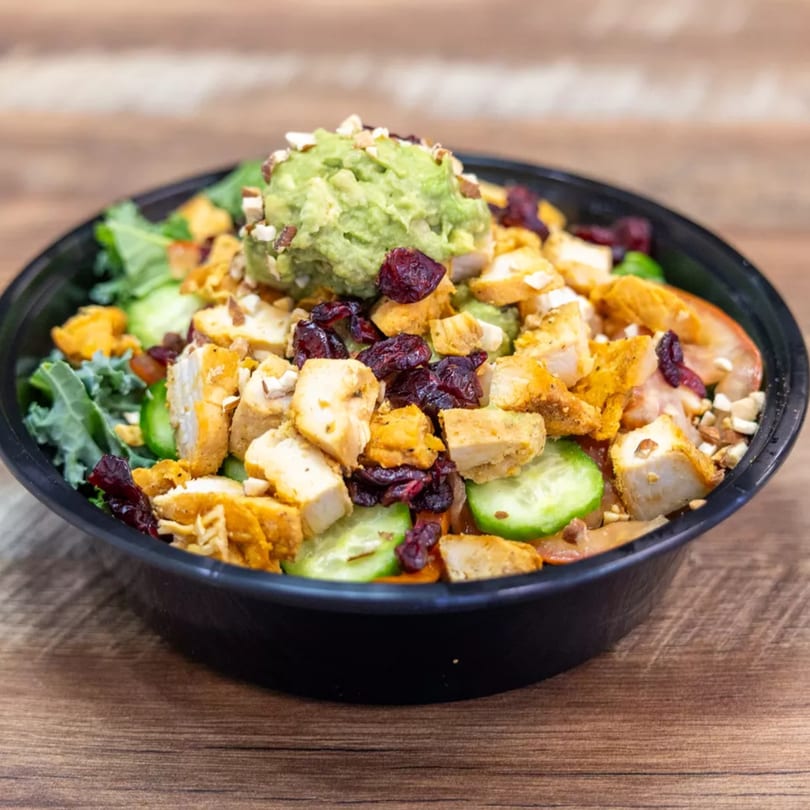 A vibrant salad with chicken, cranberries, cucumbers, and guacamole in a black bowl on a wooden table.