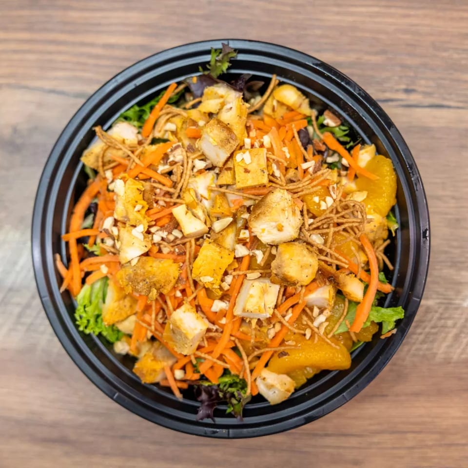Overhead view of a vibrant salad with chicken, carrots, oranges, and crispy noodles in a black bowl.