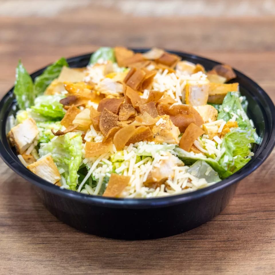 A Caesar salad with chicken, shredded cheese, and crispy pita bread in a black bowl.