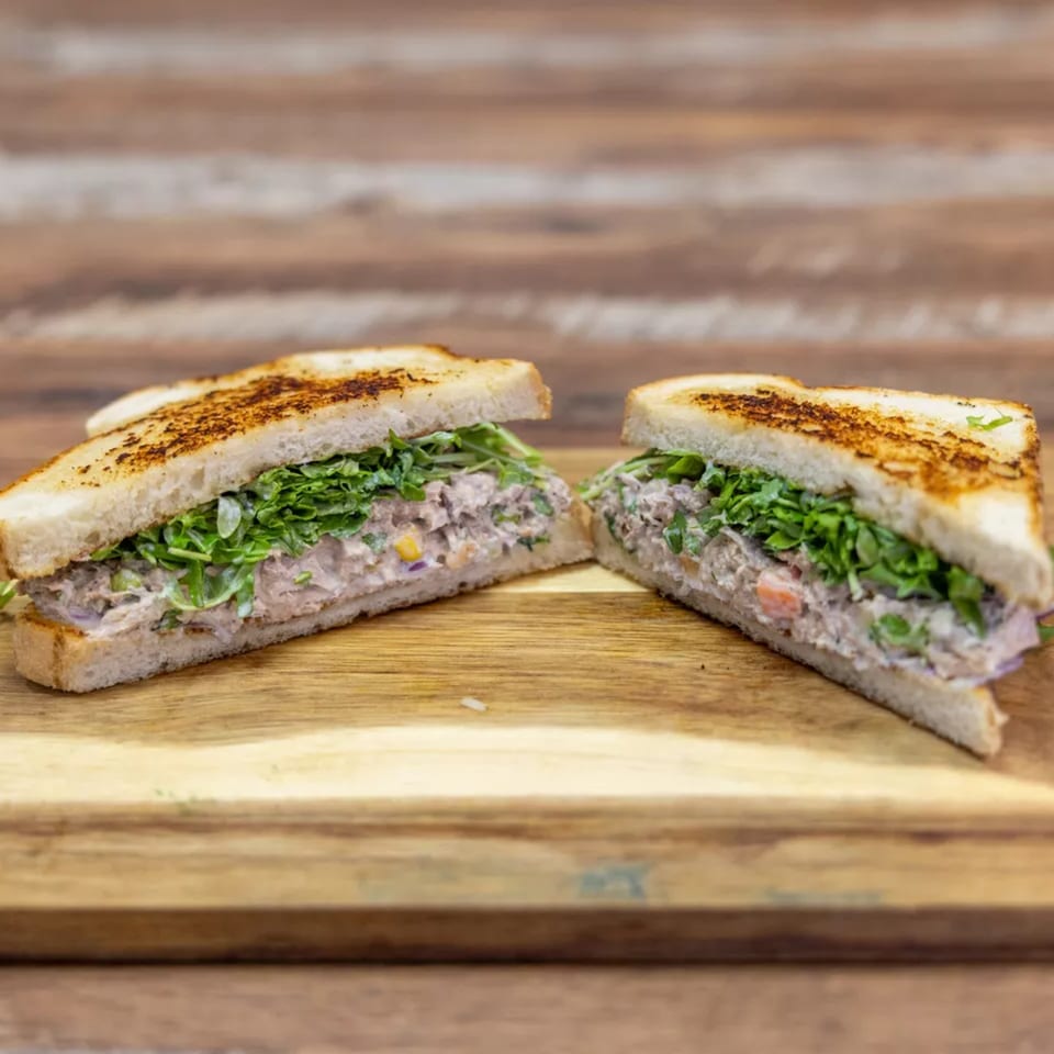 Two halves of a tuna salad sandwich with greens on toasted bread, on a wooden board.