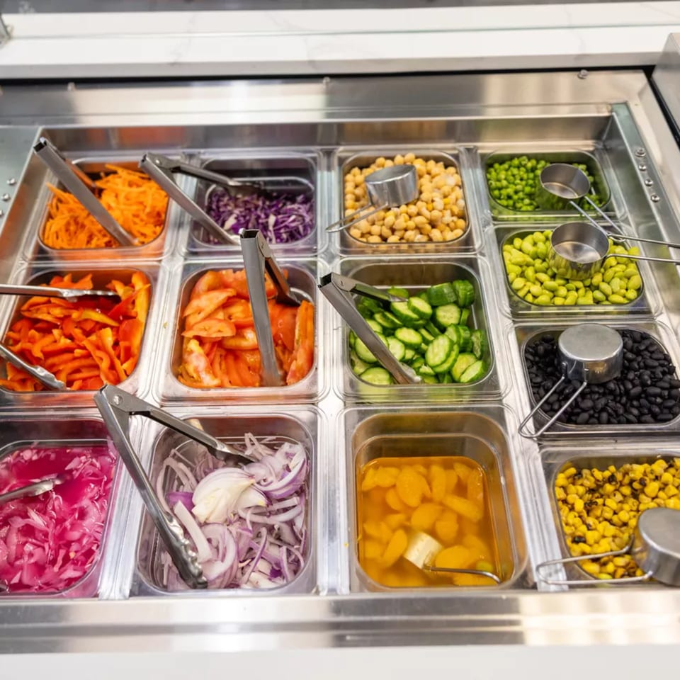 A salad bar with various fresh toppings like carrots, tomatoes, cucumbers, chickpeas, and beans.